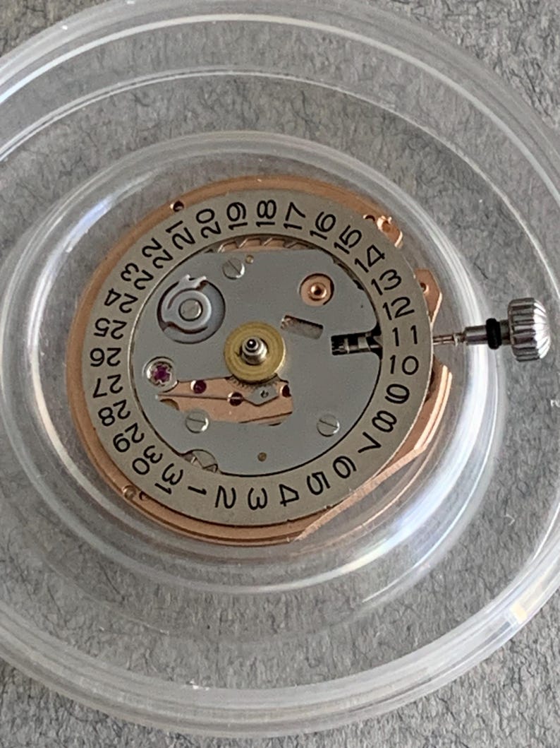 Puede incluir: Primer plano de un movimiento de reloj con una esfera plateada y dorada. La esfera presenta n&uacute;meros negros que indican la fecha y la hora. Se puede ver una peque&ntilde;a corona y otros componentes mec&aacute;nicos. El movimiento est&aacute; encerrado en un soporte de pl&aacute;stico transparente.