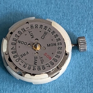 May include: A disassembled watch movement with a silver dial and white plastic casing. The dial displays the days of the week in English and German, along with the date. A small crown is attached to the side.