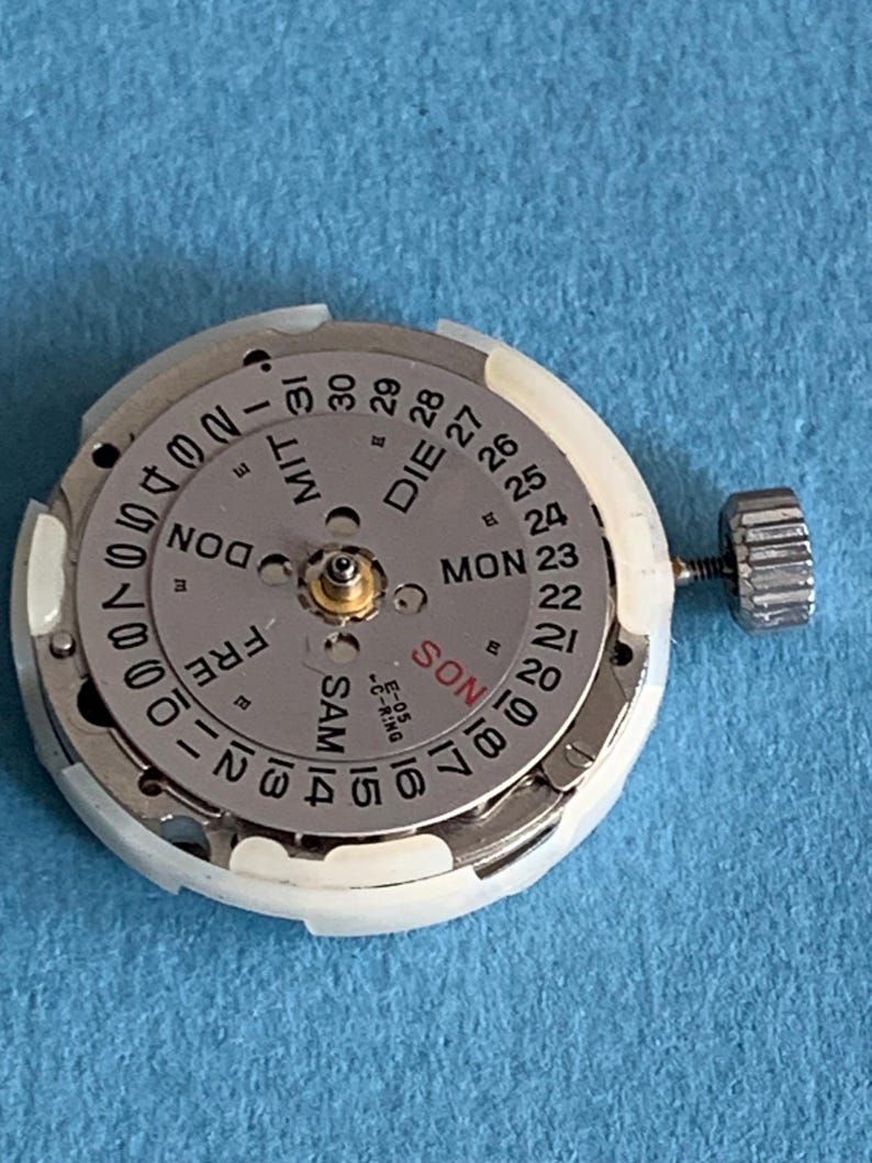 May include: A disassembled watch movement with a silver dial and white plastic casing. The dial displays the days of the week in English and German, along with the date. A small crown is attached to the side.