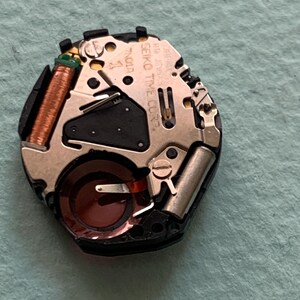 May include: A detailed view of a watch movement, showcasing small gears, coils, and a battery. The movement is mainly silver and black, with copper-coloured coils and a dark red component. The text "SEIKO TIME CORP" is visible.