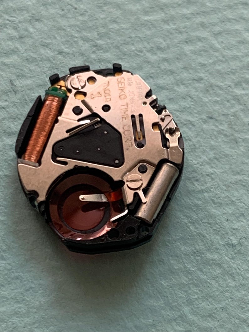 May include: A detailed view of a watch movement, showcasing small gears, coils, and a battery. The movement is mainly silver and black, with copper-coloured coils and a dark red component. The text "SEIKO TIME CORP" is visible.