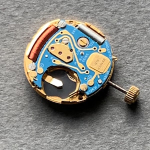 May include: Close-up of a watch movement with a gold-colored frame and a blue circuit board. The movement features a small screw, a coil, and the text "ZENITH" and "SWISS". The watch movement has seven jewels.