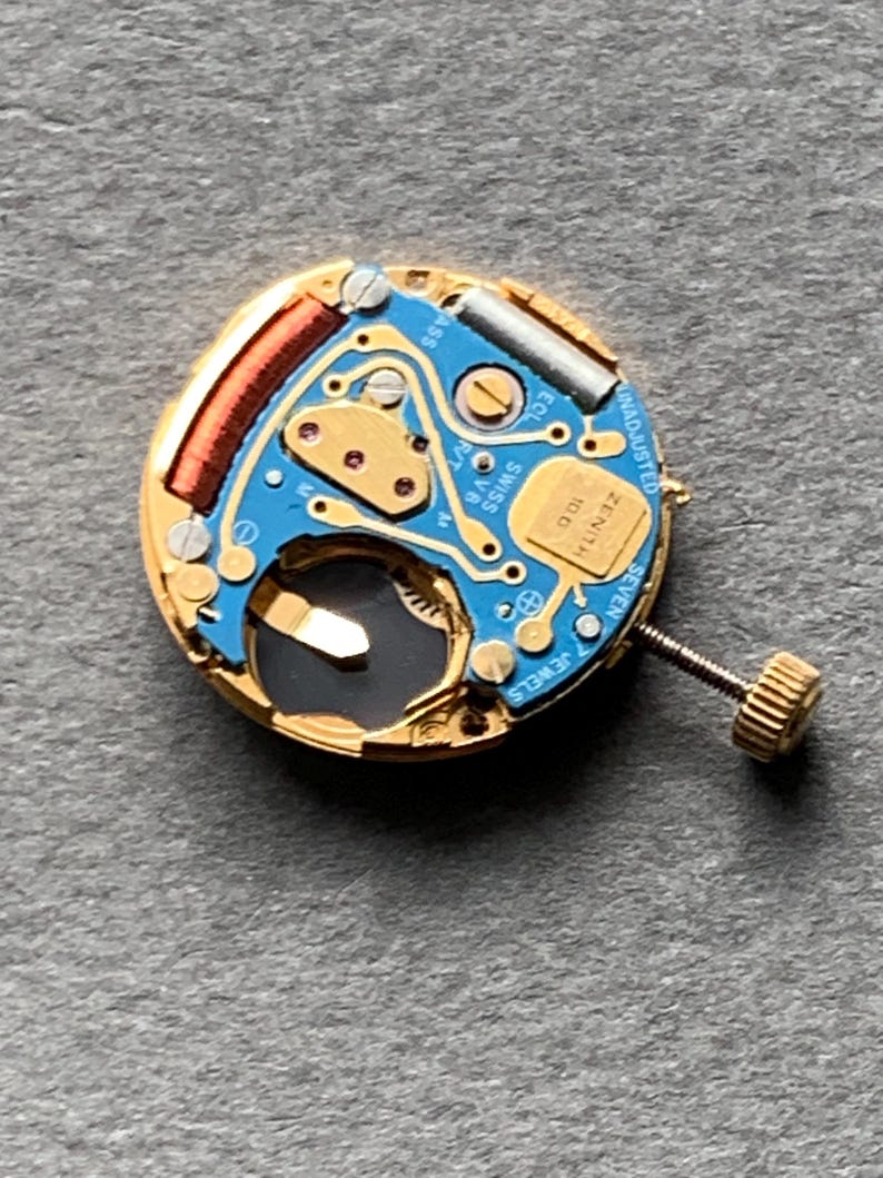 May include: Close-up of a watch movement with a gold-colored frame and a blue circuit board. The movement features a small screw, a coil, and the text "ZENITH" and "SWISS". The watch movement has seven jewels.