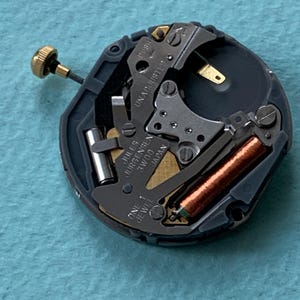 K&ouml;nnte beinhalten: Nahaufnahme eines Uhrwerks mit dem Text "Jules Jurgensen 3W00 Japan". Der Mechanismus hat einen goldfarbenen Stiel und eine kupferfarbene Spule. Die Worte "ONE 1 JEWEL" sind ebenfalls sichtbar.