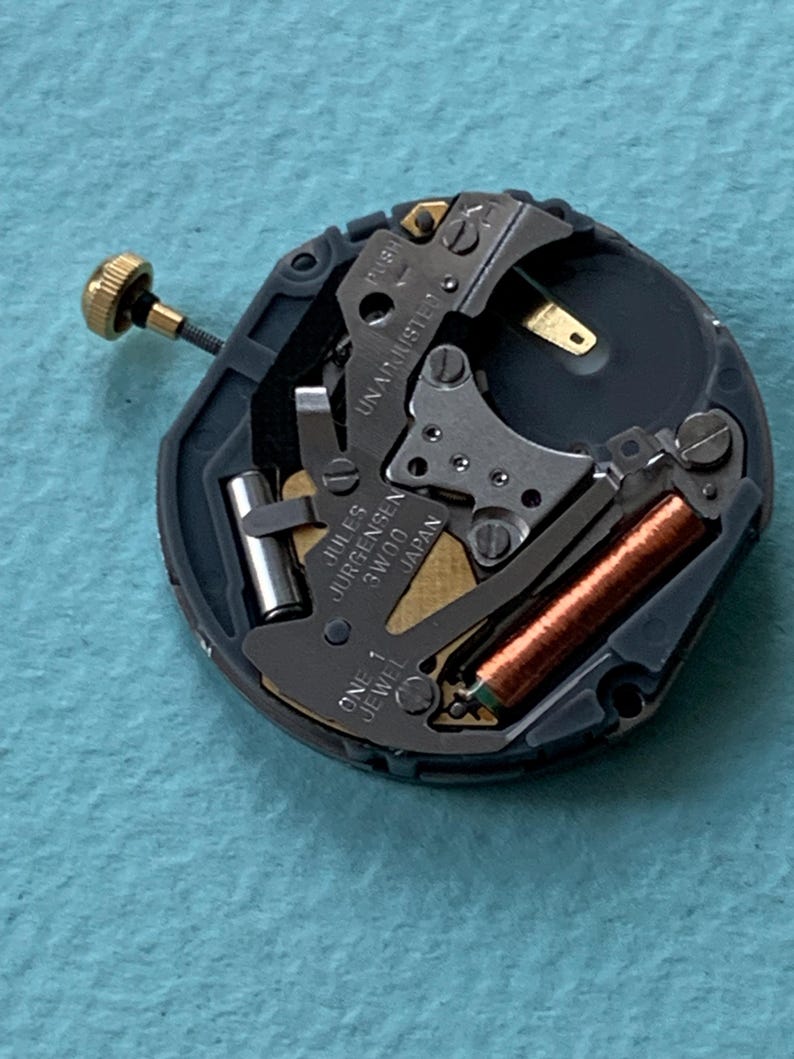 K&ouml;nnte beinhalten: Nahaufnahme eines Uhrwerks mit dem Text "Jules Jurgensen 3W00 Japan". Der Mechanismus hat einen goldfarbenen Stiel und eine kupferfarbene Spule. Die Worte "ONE 1 JEWEL" sind ebenfalls sichtbar.