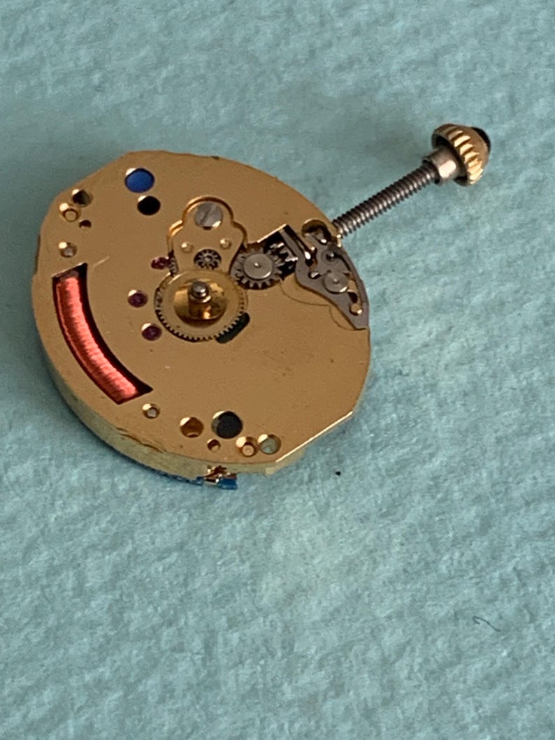May include: A gold-toned watch movement with intricate gears, a red coil, and a screw-down crown. The watch mechanism is oval-shaped and features small jewels and various components. The background is a light blue surface.