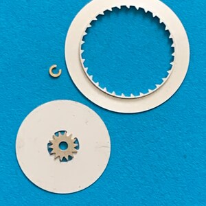 May include: A collection of watch parts, including two white circular pieces with toothed edges, a small gear, and a tiny gold-colored ring. The parts are arranged on a blue surface, suggesting a repair or assembly context.