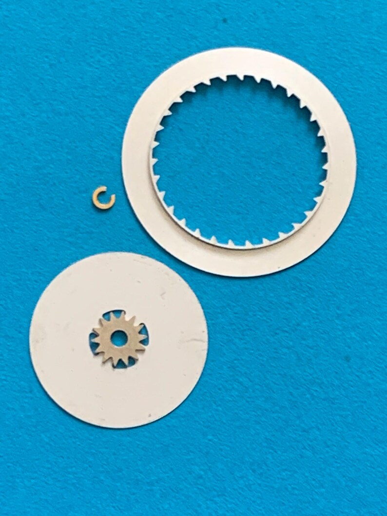 May include: A collection of watch parts, including two white circular pieces with toothed edges, a small gear, and a tiny gold-colored ring. The parts are arranged on a blue surface, suggesting a repair or assembly context.