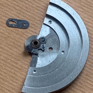 Pu&ograve; includere: Un movimento di orologio smontato, con un grande componente metallico semicircolare con scanalature circolari e diversi piccoli ingranaggi. &Egrave; presente anche un piccolo pezzo di metallo di colore scuro. I componenti sono esposti su una superficie di legno.