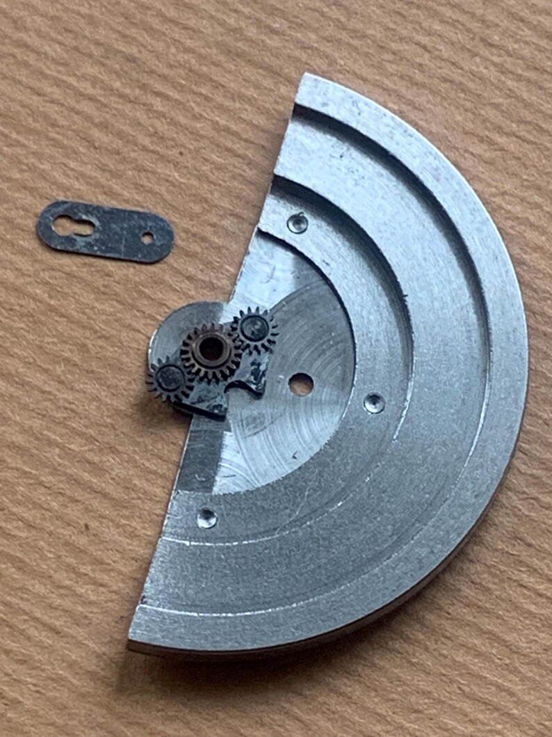 Pu&ograve; includere: Un movimento di orologio smontato, con un grande componente metallico semicircolare con scanalature circolari e diversi piccoli ingranaggi. &Egrave; presente anche un piccolo pezzo di metallo di colore scuro. I componenti sono esposti su una superficie di legno.