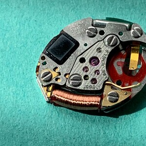 May include: Close-up of a Seiko watch movement with intricate gears, screws, and a red component. The text "SEIKO TIME CORP" and "JAPAN" are visible. The watch mechanism is set against a teal background.