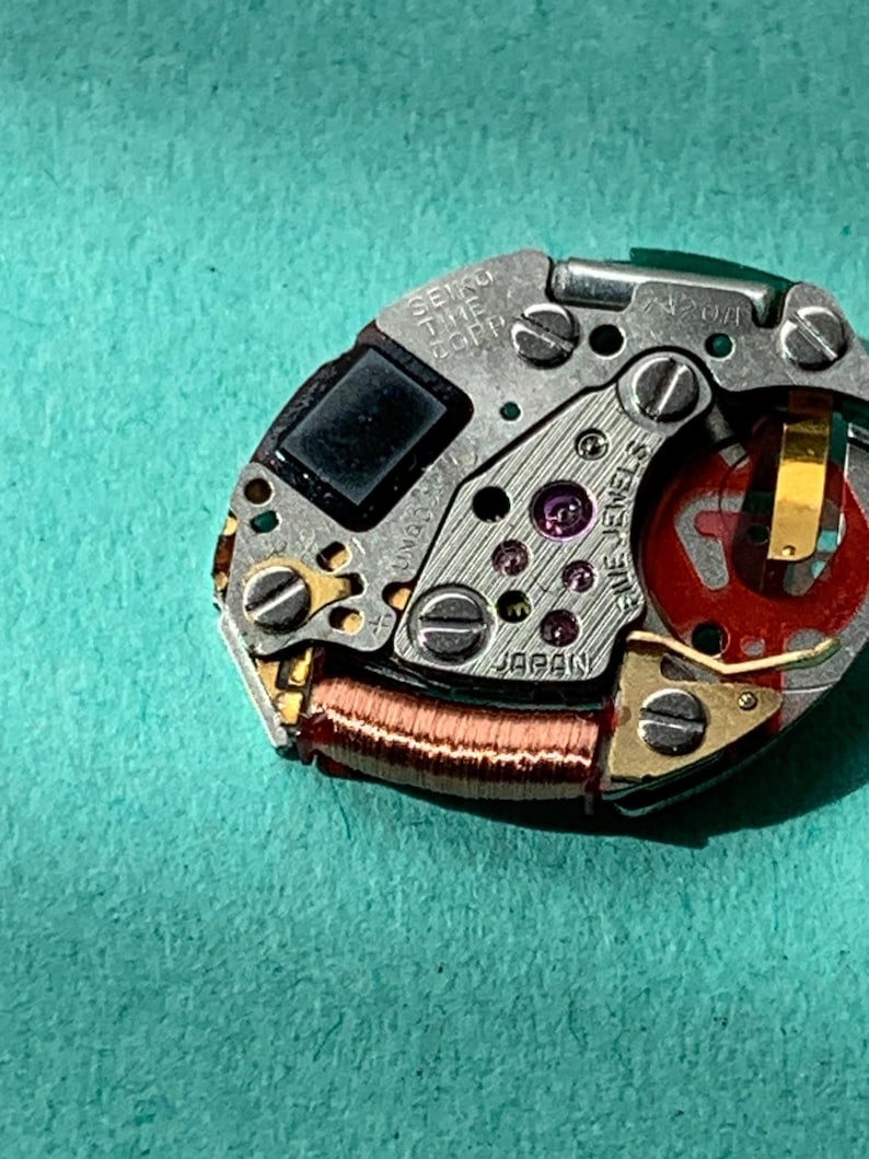 May include: Close-up of a Seiko watch movement with intricate gears, screws, and a red component. The text "SEIKO TIME CORP" and "JAPAN" are visible. The watch mechanism is set against a teal background.