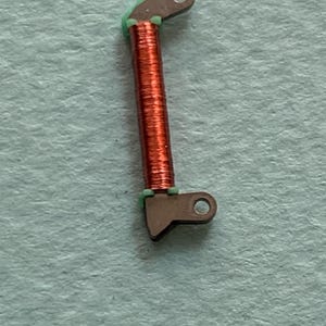 May include: Close-up of a small, mechanical component. The component features a copper-colored coil wrapped around a central core, with dark gray metal brackets at each end. Green material is visible between the coil and the brackets. The background is a light blue-green.