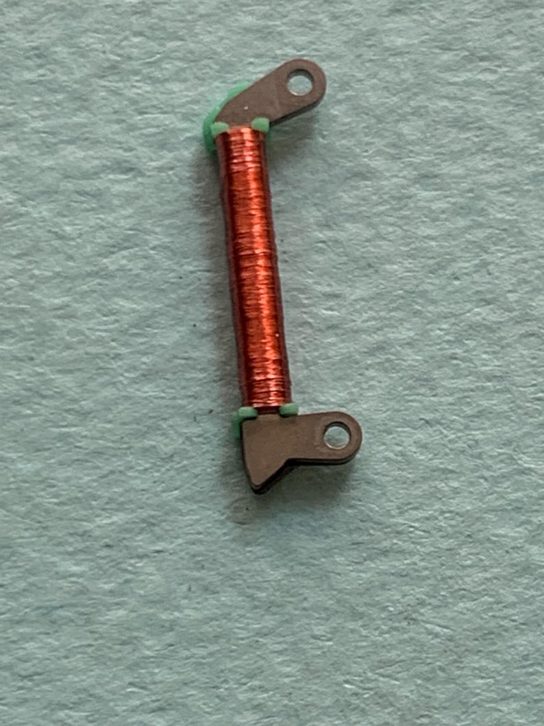 May include: Close-up of a small, mechanical component. The component features a copper-colored coil wrapped around a central core, with dark gray metal brackets at each end. Green material is visible between the coil and the brackets. The background is a light blue-green.