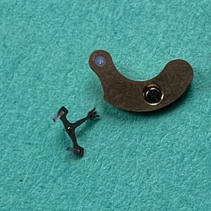 May include: Two small watch parts on a teal surface. One is a curved, brown metal piece with a circular hole and a small blue dot. The other is a small, black metal piece with a Y-shape.