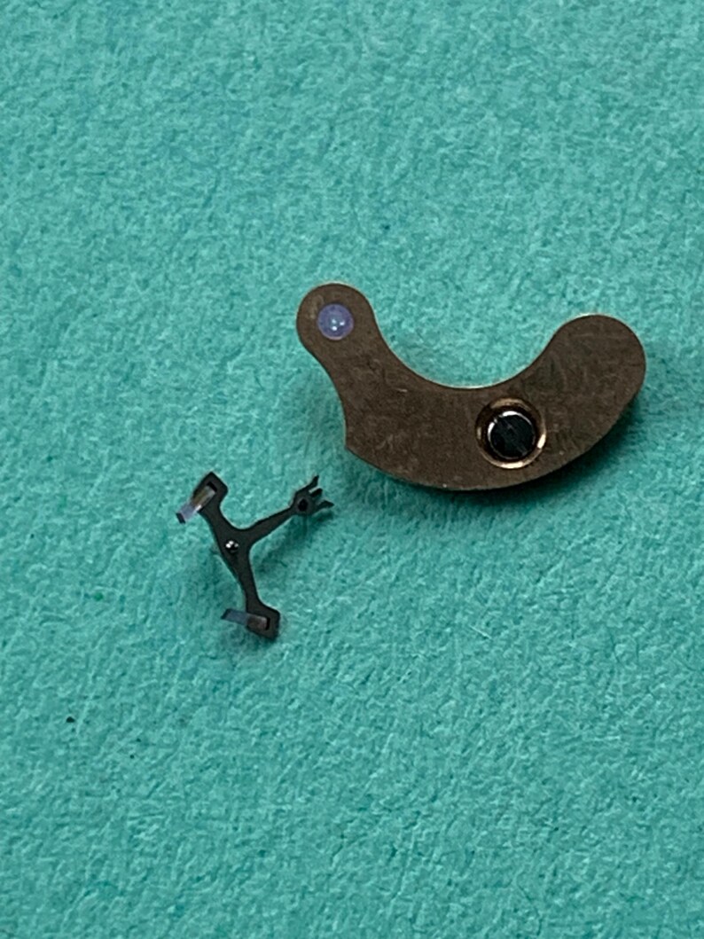 May include: Two small watch parts on a teal surface. One is a curved, brown metal piece with a circular hole and a small blue dot. The other is a small, black metal piece with a Y-shape.