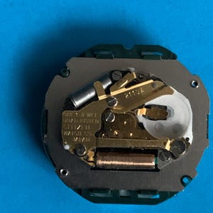 May include: A Citizen watch movement, with gold-coloured gears and components, set on a grey base with a green border. The mechanism has text that reads "QUARTZ 1 JEWEL ADJUSTED CITIZEN WATCH CO. JAPAN".