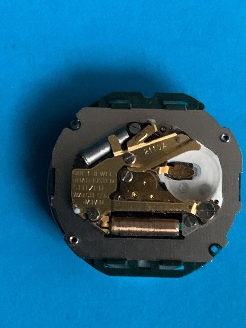 May include: A Citizen watch movement, with gold-coloured gears and components, set on a grey base with a green border. The mechanism has text that reads "QUARTZ 1 JEWEL ADJUSTED CITIZEN WATCH CO. JAPAN".