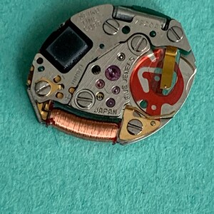 May include: A close-up of a Seiko watch movement. The silver-toned mechanism features small screws, a copper coil, and red and purple jewels. The text "SEIKO TIME CORP" and "JAPAN" are visible.