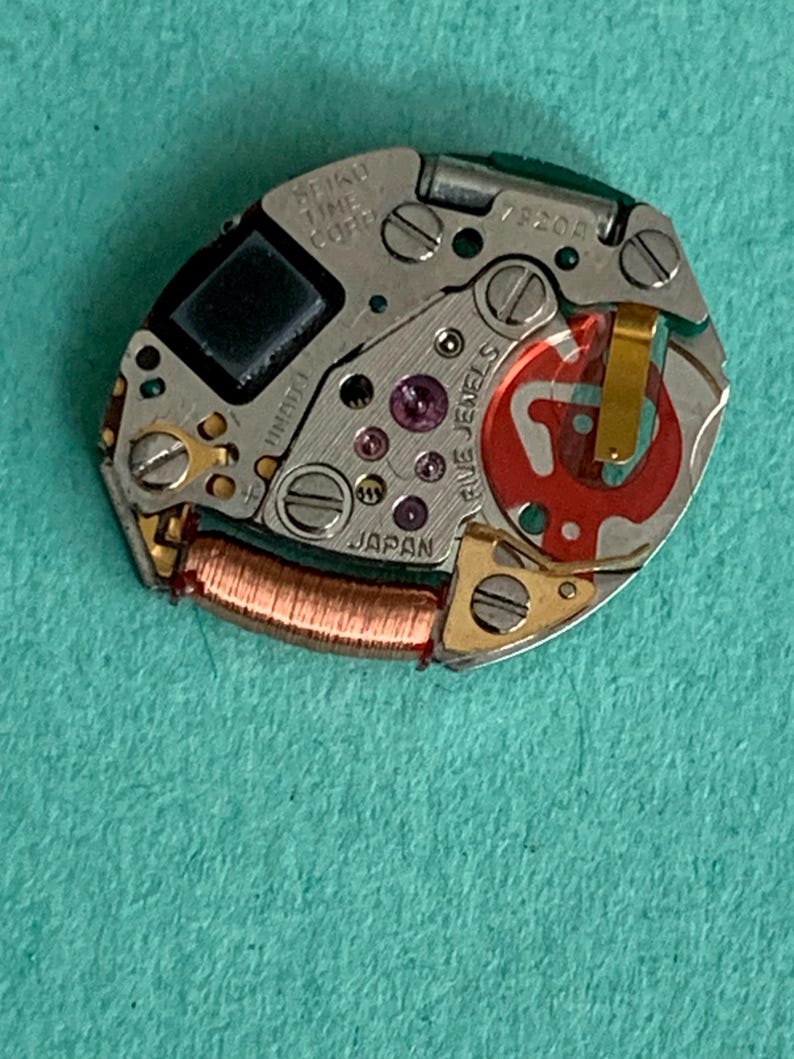 May include: A close-up of a Seiko watch movement. The silver-toned mechanism features small screws, a copper coil, and red and purple jewels. The text "SEIKO TIME CORP" and "JAPAN" are visible.