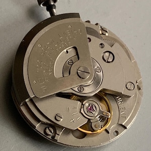 May include: A silver-toned mechanical watch movement featuring a winding stem at the top. The metal plate is engraved with "ORIENT WATCH CO., LTD.", "TWENTY-ONE JEWELS", and "JAPAN 48948". A gold-colored balance wheel with a purple jewel is visible.