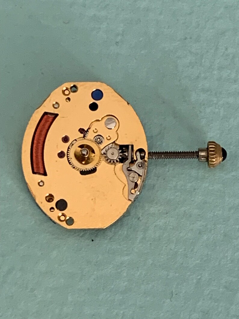 May include: A close-up of a watch movement with gold-colored components. The movement features gears, a screw, and a crown. A red-brown curved piece is on the left side. The background is a light blue-green color.