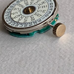 Puede incluir: Un movimiento de reloj con una esfera blanca que muestra los d&iacute;as de la semana y las fechas numeradas. La esfera est&aacute; rodeada por un anillo de metal plateado. Una corona dorada est&aacute; unida al lateral. El movimiento se asienta sobre una base verde.