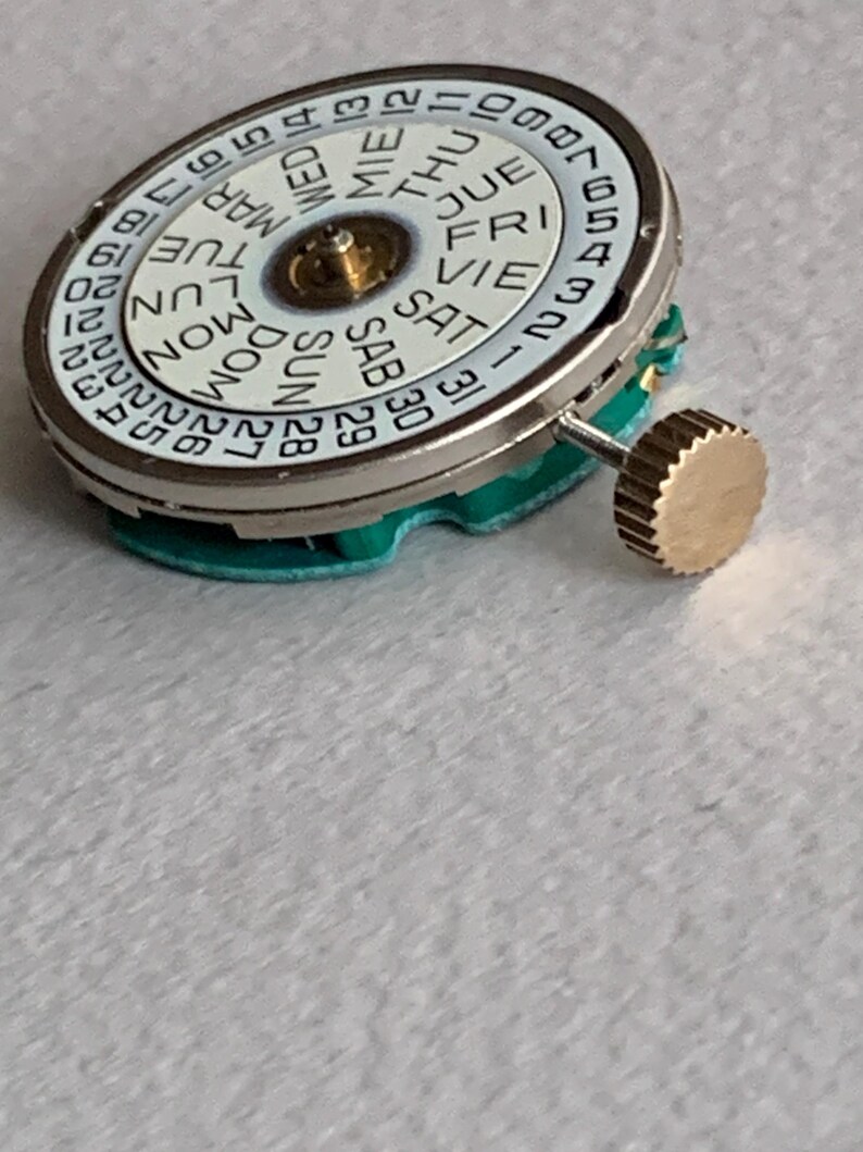 Puede incluir: Un movimiento de reloj con una esfera blanca que muestra los d&iacute;as de la semana y las fechas numeradas. La esfera est&aacute; rodeada por un anillo de metal plateado. Una corona dorada est&aacute; unida al lateral. El movimiento se asienta sobre una base verde.