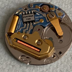 Może przedstawiać: Zbliżenie mechanizmu zegarka z misternymi detalami. Mechanizm zegarka zawiera złote, srebrne i niebieskie elementy, w tym płytkę obwodu z napisem "CERTINA 729 SWISS V8". Zegarek ma r&oacute;wnież napis "SEVEN 7 JEWELS UNADJUSTED".