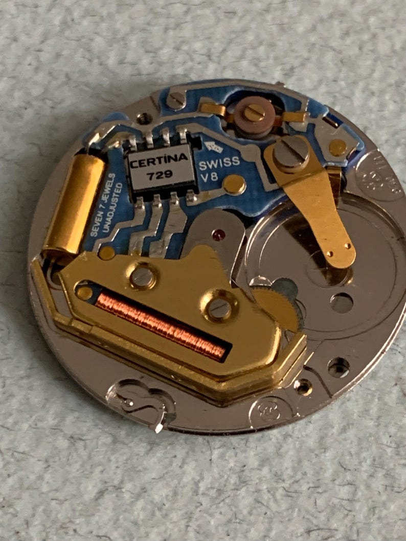 Może przedstawiać: Zbliżenie mechanizmu zegarka z misternymi detalami. Mechanizm zegarka zawiera złote, srebrne i niebieskie elementy, w tym płytkę obwodu z napisem "CERTINA 729 SWISS V8". Zegarek ma r&oacute;wnież napis "SEVEN 7 JEWELS UNADJUSTED".