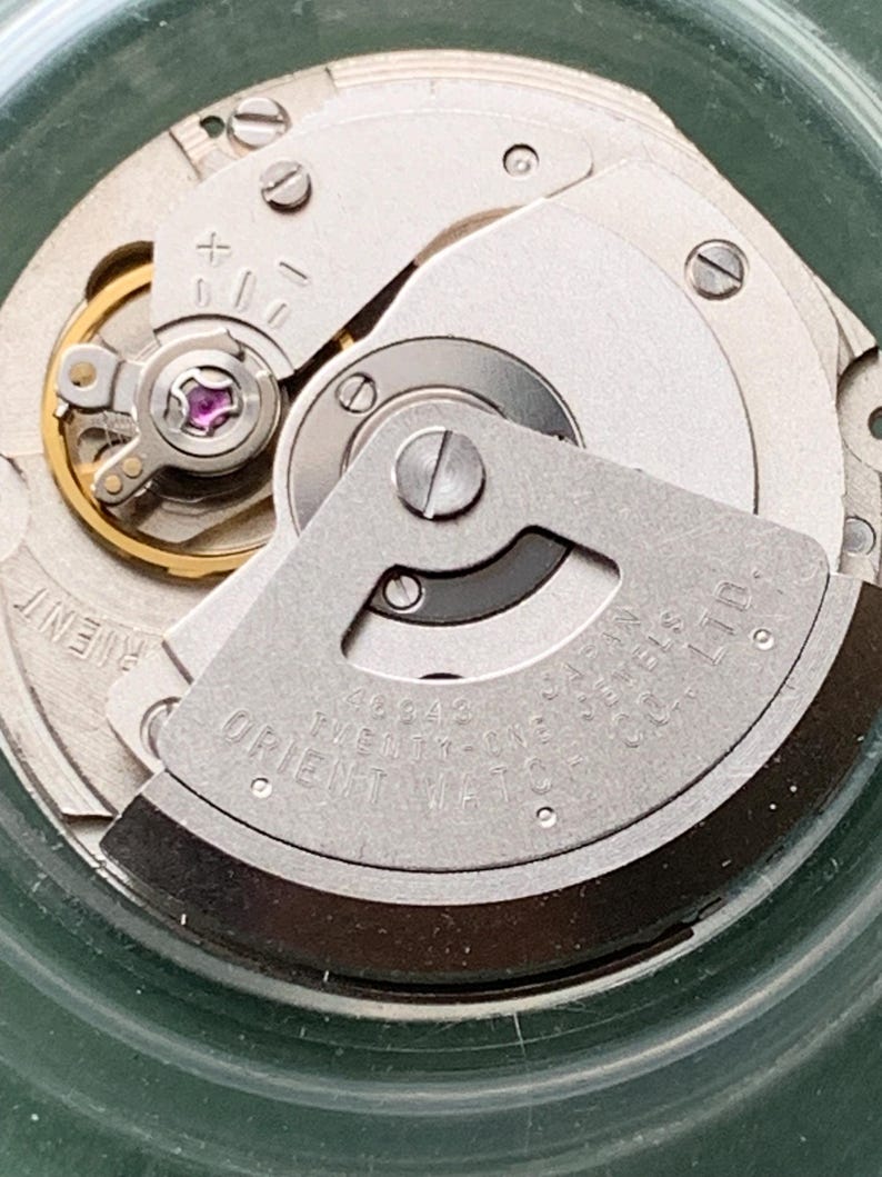 May include: Close-up of a watch movement with silver gears and components. The text "ORIENT WATCH CO. LTD." is visible. The movement features a jewel-toned purple stone and various screws.