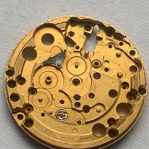 May include: Close-up of a gold-colored watch movement, showcasing intricate gears, holes, and circular components. The mechanical parts are arranged in a circular pattern, with a few small, colored jewels visible.