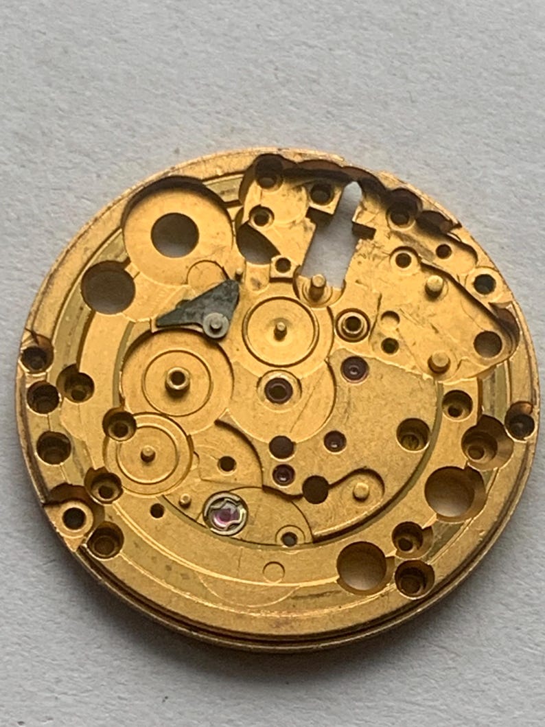May include: Close-up of a gold-colored watch movement, showcasing intricate gears, holes, and circular components. The mechanical parts are arranged in a circular pattern, with a few small, colored jewels visible.