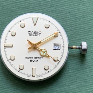 K&ouml;nnte beinhalten: Ein Casio Quartz Zifferblatt mit cremefarbenem Zifferblatt, goldenen Zeigern und Markierungen. Das Zifferblatt zeigt das Datum an und ist mit "WATER RESIST 50M" gekennzeichnet. Die Uhr hat eine silberne Krone und ist vor einem gr&uuml;nen Hintergrund platziert.