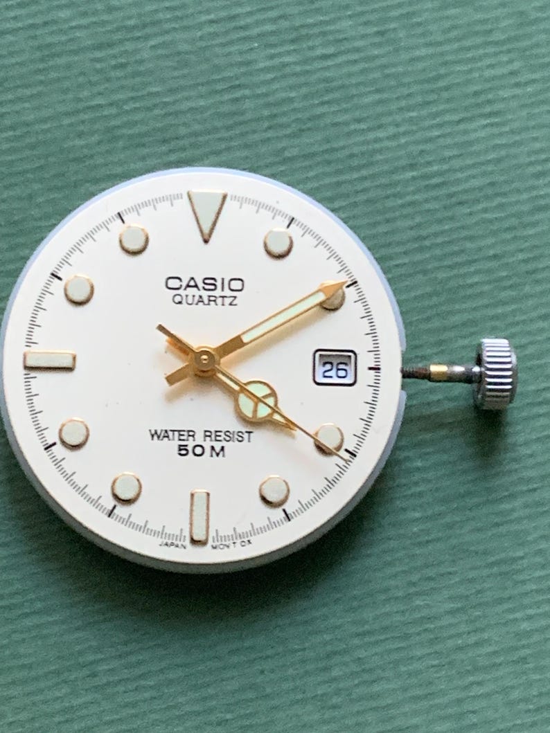 K&ouml;nnte beinhalten: Ein Casio Quartz Zifferblatt mit cremefarbenem Zifferblatt, goldenen Zeigern und Markierungen. Das Zifferblatt zeigt das Datum an und ist mit "WATER RESIST 50M" gekennzeichnet. Die Uhr hat eine silberne Krone und ist vor einem gr&uuml;nen Hintergrund platziert.