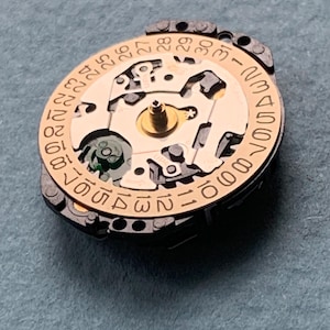 K&ouml;nnte beinhalten: Nahaufnahme eines Uhrwerks mit goldfarbenem Zentrum und kreisf&ouml;rmigem Datumszifferblatt. Das Zifferblatt zeigt Zahlen von 1 bis 31. Das Uhrwerk hat komplizierte mechanische Komponenten. Der Hintergrund ist grau.