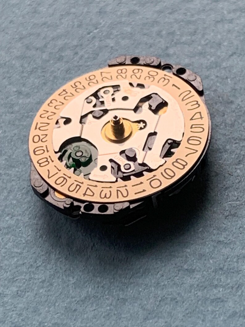K&ouml;nnte beinhalten: Nahaufnahme eines Uhrwerks mit goldfarbenem Zentrum und kreisf&ouml;rmigem Datumszifferblatt. Das Zifferblatt zeigt Zahlen von 1 bis 31. Das Uhrwerk hat komplizierte mechanische Komponenten. Der Hintergrund ist grau.