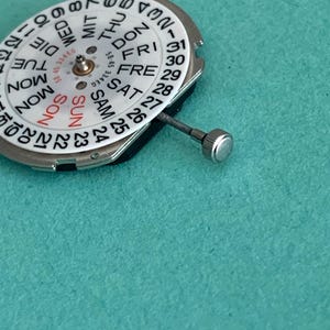 May include: A watch movement with a white dial showing days and dates. The dial has black numerals and text, with "SUN" in red. A silver stem and crown are attached. The background is teal.