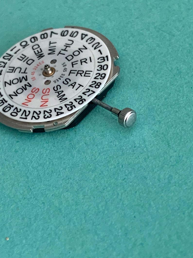May include: A watch movement with a white dial showing days and dates. The dial has black numerals and text, with "SUN" in red. A silver stem and crown are attached. The background is teal.