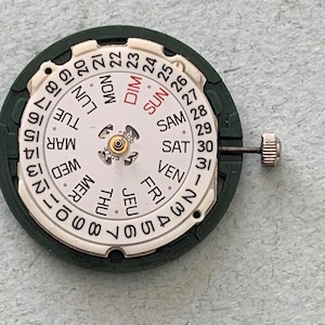 May include: A watch movement with a white dial displaying the days of the week in English and numbers 1-31. The dial is surrounded by a dark green ring. A small silver crown is attached to the side.