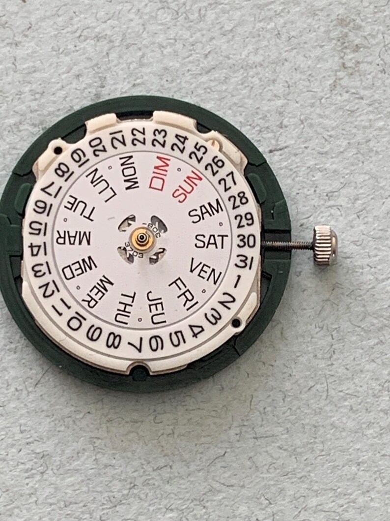 May include: A watch movement with a white dial displaying the days of the week in English and numbers 1-31. The dial is surrounded by a dark green ring. A small silver crown is attached to the side.
