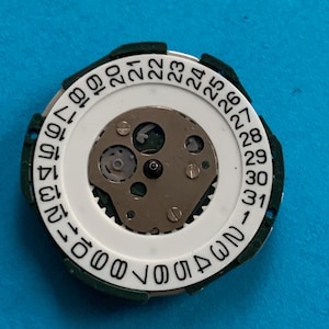 May include: A watch movement component with a white circular dial featuring black numbers from 1 to 31, indicating the days of the month. The dial is surrounded by a green plastic ring. The centre reveals the intricate mechanical workings of the watch.