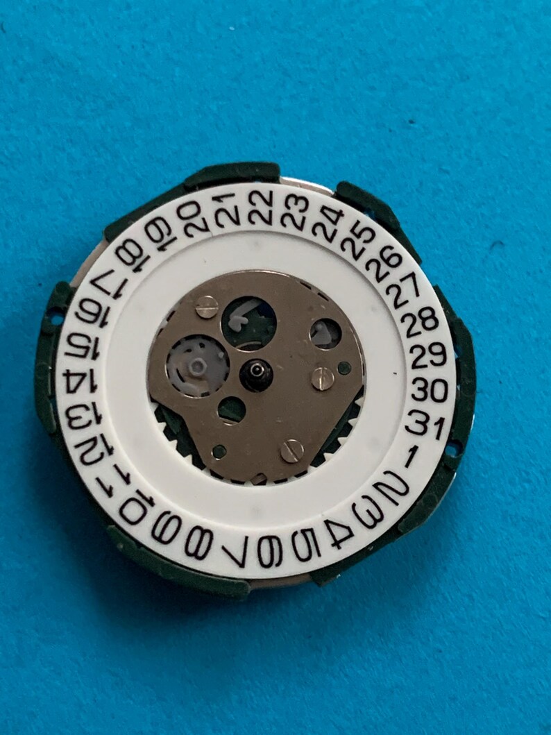 May include: A watch movement component with a white circular dial featuring black numbers from 1 to 31, indicating the days of the month. The dial is surrounded by a green plastic ring. The centre reveals the intricate mechanical workings of the watch.