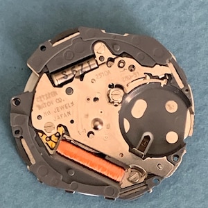May include: Close-up of a Citizen watch movement with the text "Citizen Watch Co. No Jewels Japan". The movement has a silver and gray metal frame with an orange coil and various small components.