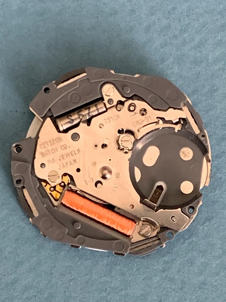 May include: Close-up of a Citizen watch movement with the text "Citizen Watch Co. No Jewels Japan". The movement has a silver and gray metal frame with an orange coil and various small components.