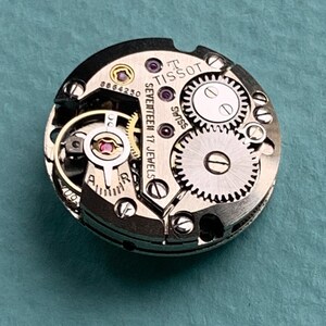 May include: Close-up of a silver Tissot watch movement with the text "Tissot Seventeen 17 Jewels Swiss" visible. The movement has a variety of gears and a red jewel.