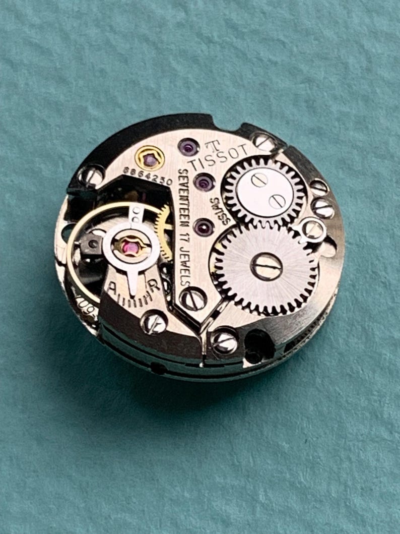 May include: Close-up of a silver Tissot watch movement with the text "Tissot Seventeen 17 Jewels Swiss" visible. The movement has a variety of gears and a red jewel.