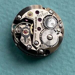 May include: Close-up of a silver Tissot watch movement with the text "SEVENTEEN 17 JEWELS" and "TISSOT" visible. The movement has a number "8864250" printed on it.