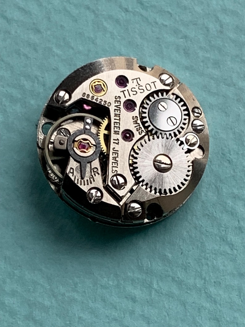 May include: Close-up of a silver Tissot watch movement with the text "SEVENTEEN 17 JEWELS" and "TISSOT" visible. The movement has a number "8864250" printed on it.