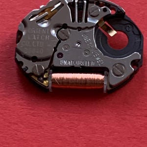 May include: Close-up of a watch movement with intricate metal components. The mechanism features the text "ORIENT LATCH CO. LTD" and "UNADJUSTED". A copper-colored coil is visible, along with a black plastic ring and a single jewel.